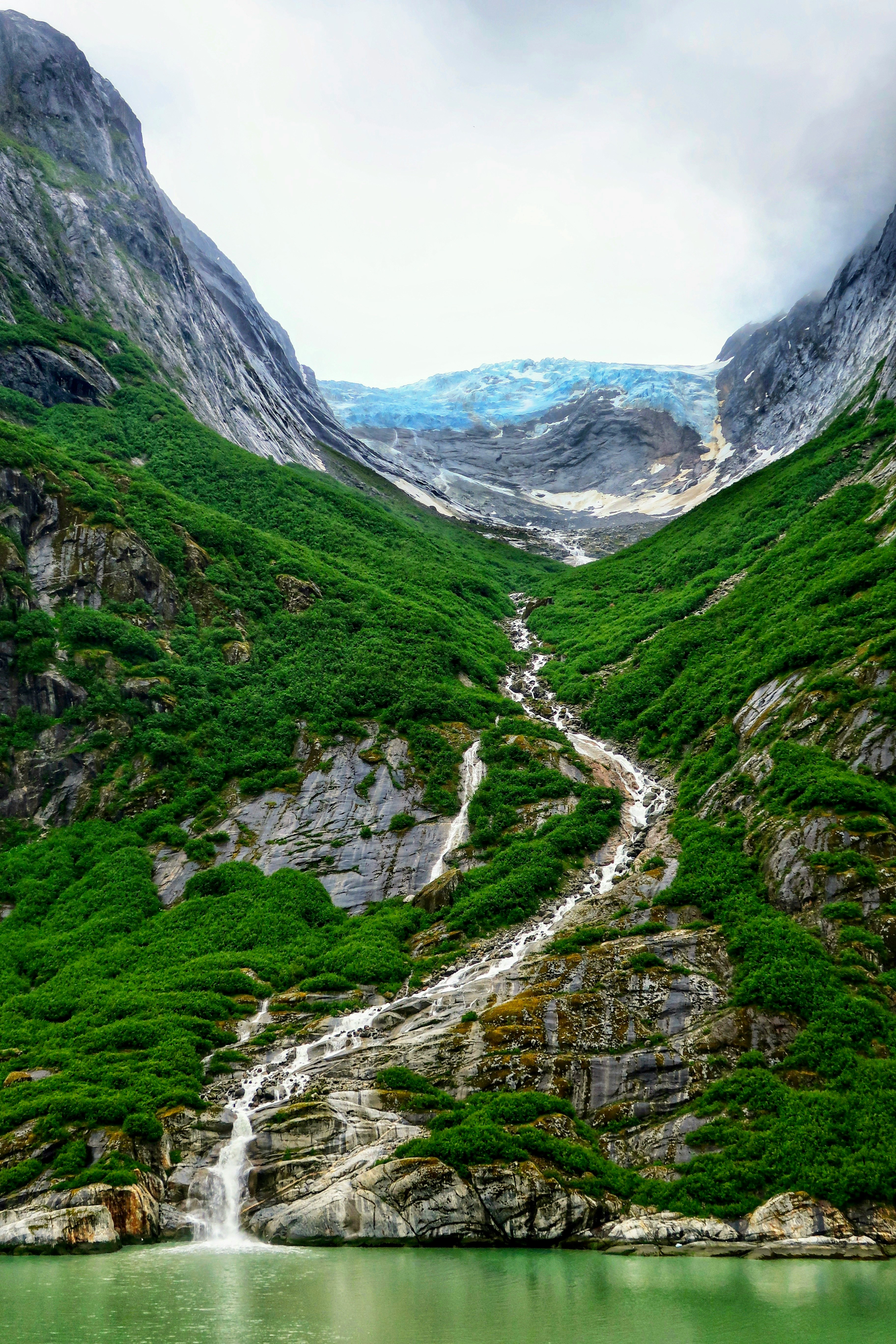A waterfall cascading through a green Alaskan fjord toward a hanging glacier