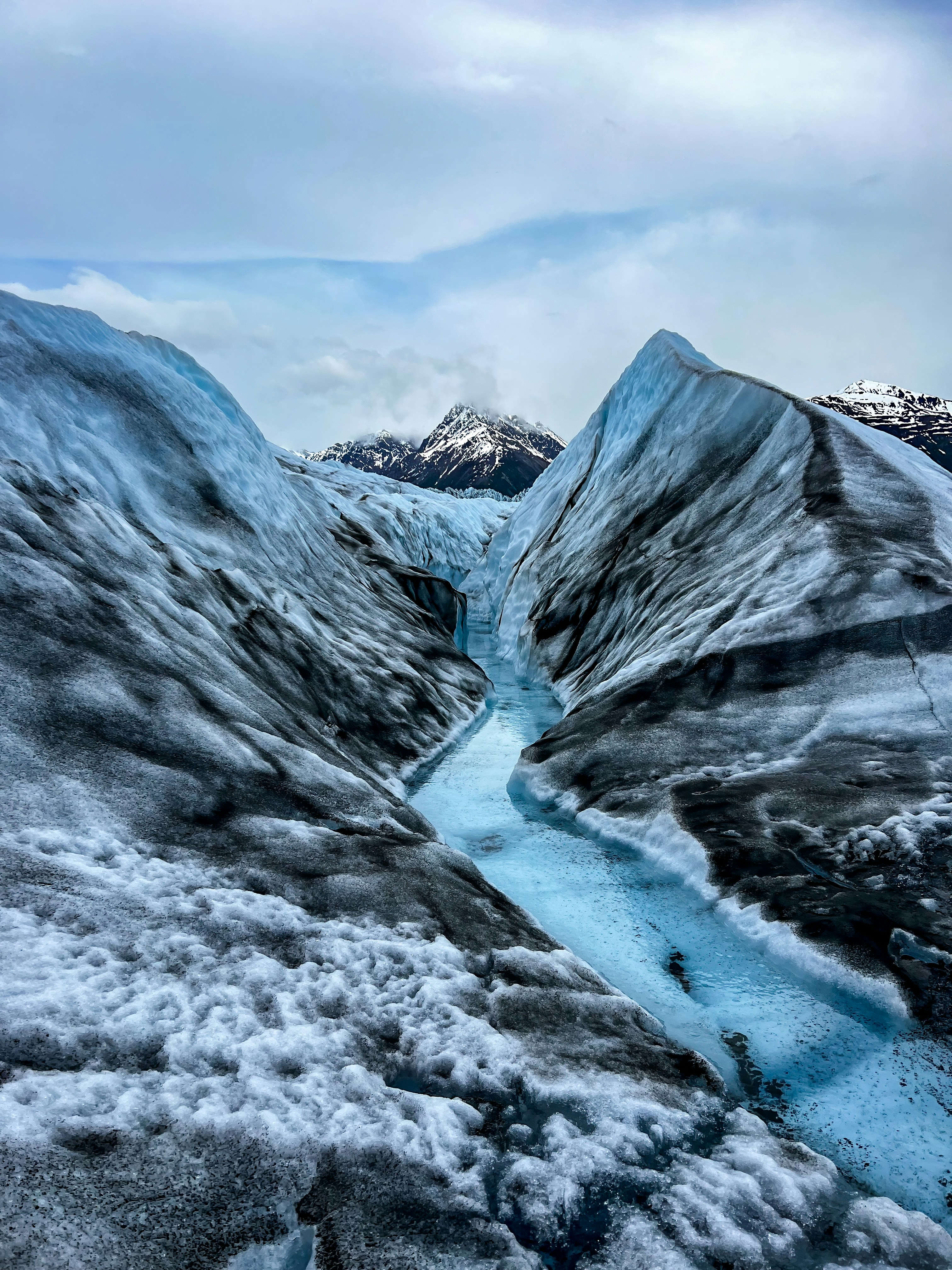 A blue meltwater channel cutting through an Alaskan glacier, mountains beyond