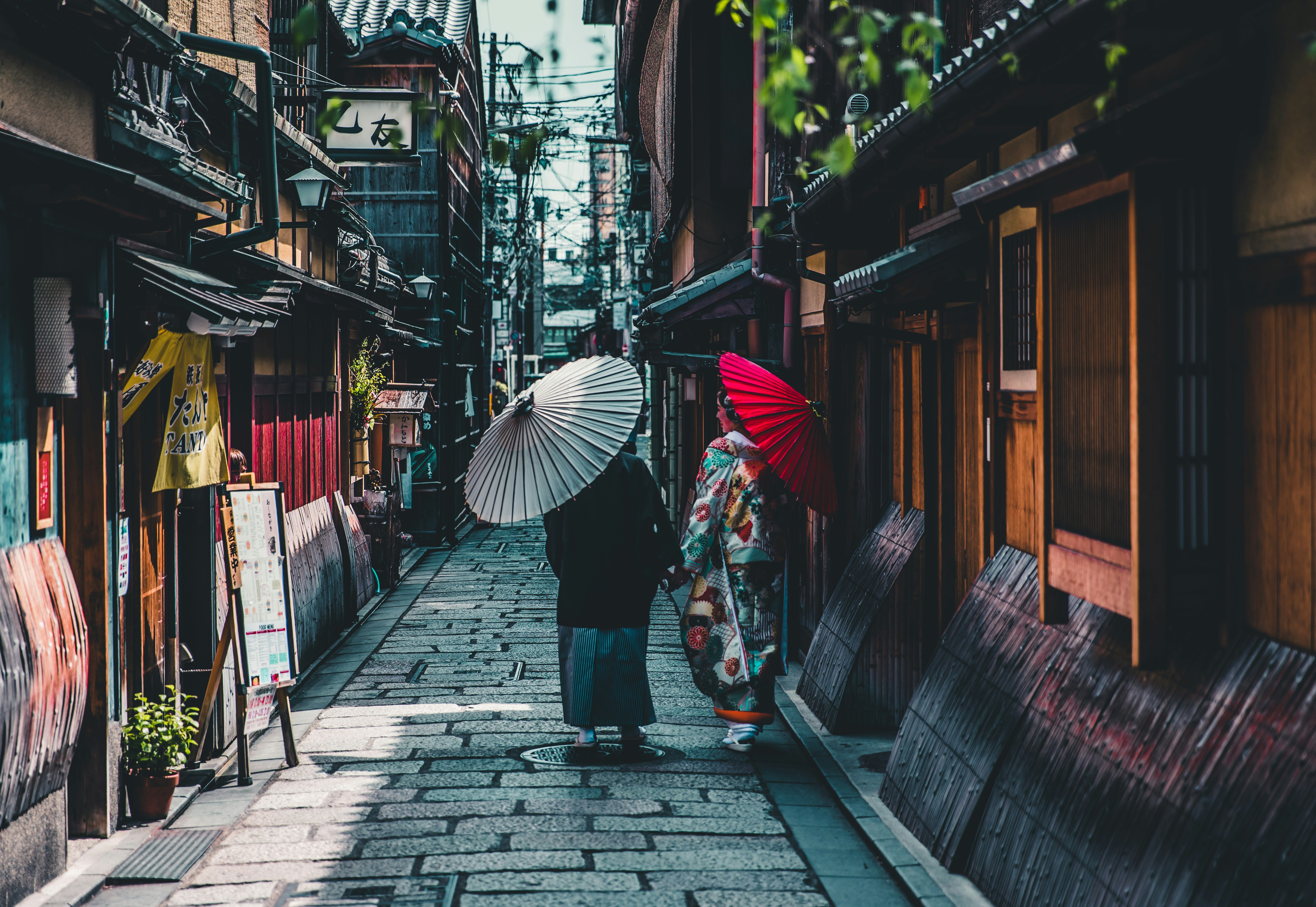 Two people in traditional dress with paper umbrellas walking a narrow Kyoto alley