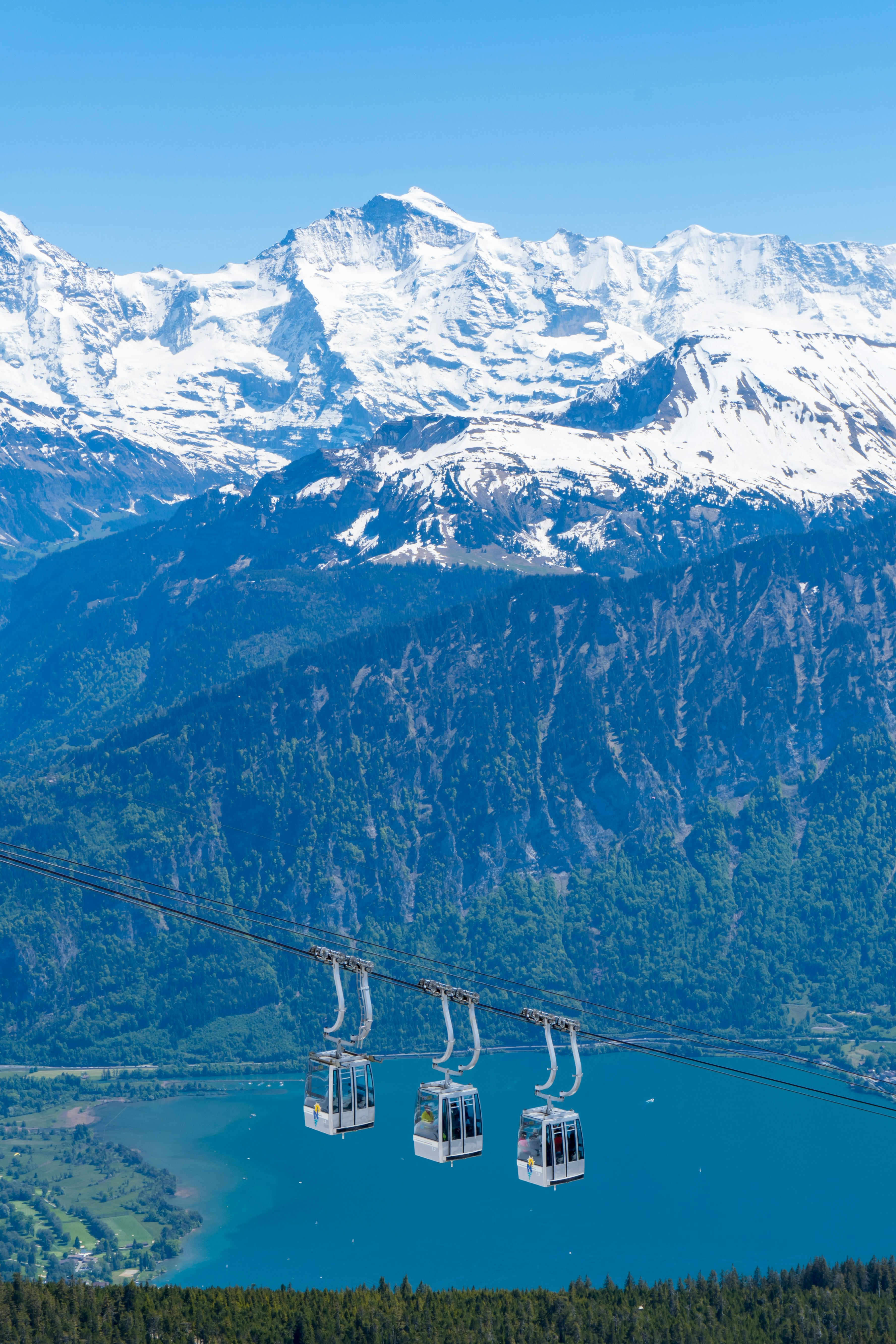 Cable cars descending toward a turquoise alpine lake with snow-capped peaks behind